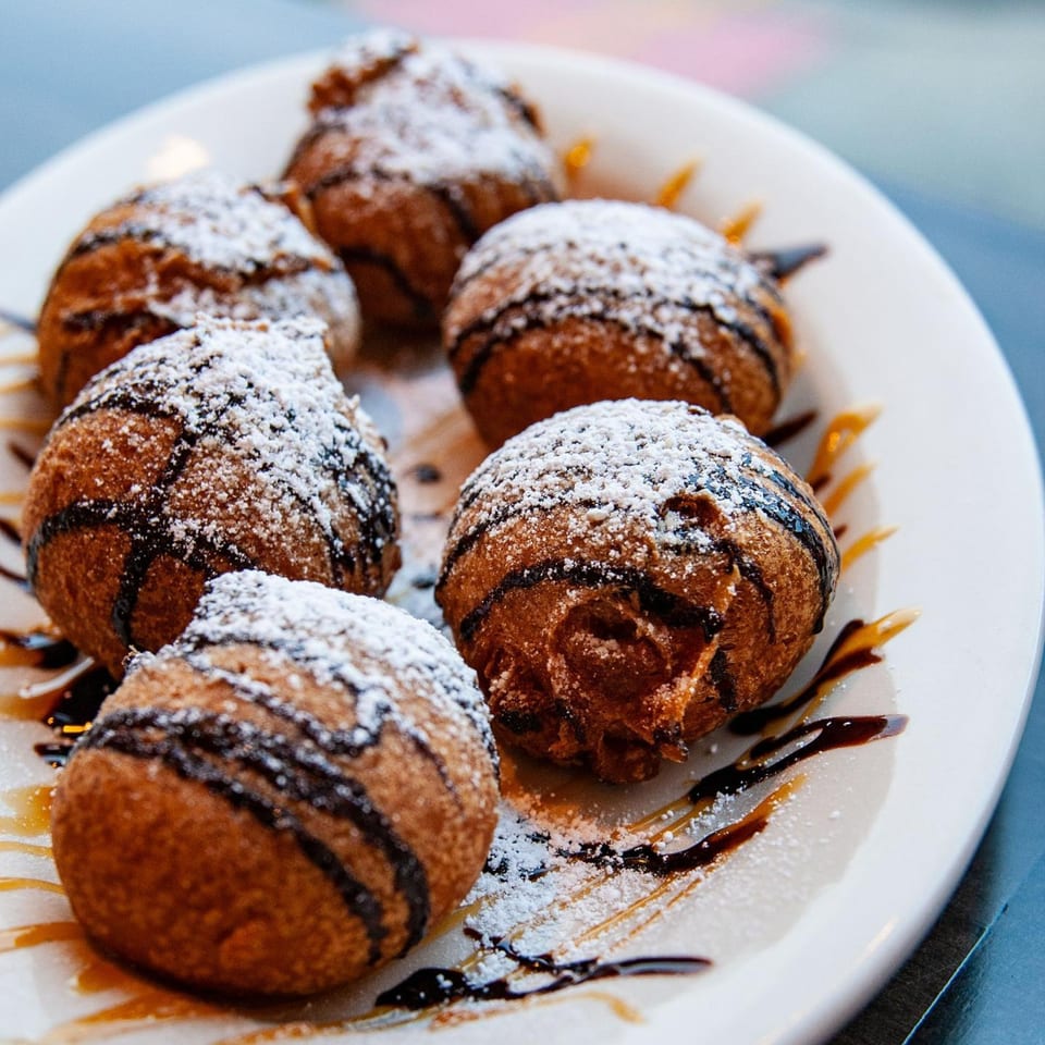 Six deep-fried breakfast pastries, drizzled with chocolate and caramel, dusted with powdered sugar.