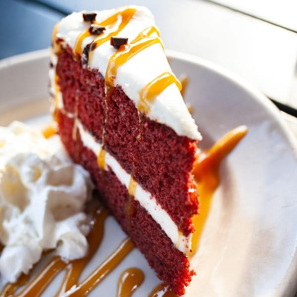 A slice of red velvet cake with white frosting, caramel drizzle, and chocolate chips, served with whipped cream.