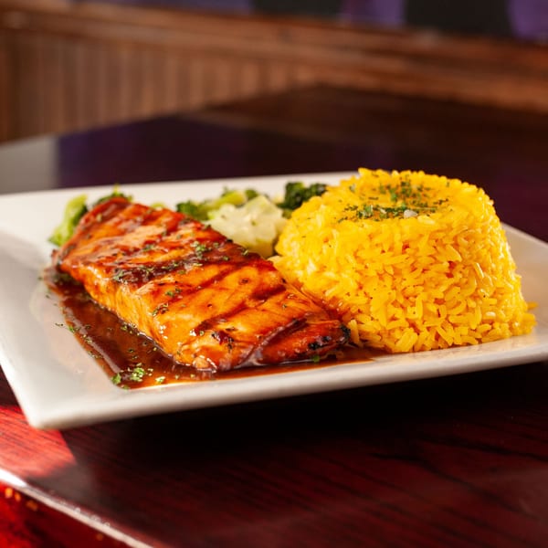 Grilled salmon with a dark glaze, yellow rice, and green vegetables on a white rectangular plate.