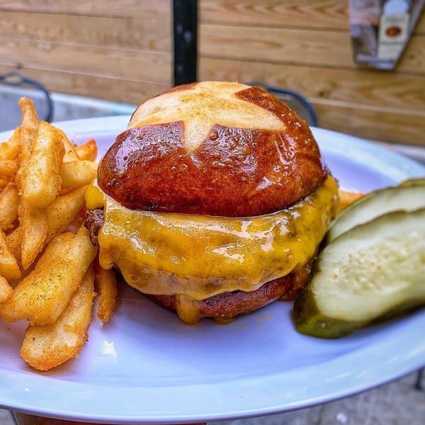 Cheeseburger on a pretzel bun with a side of seasoned fries and pickle slices on a white plate.