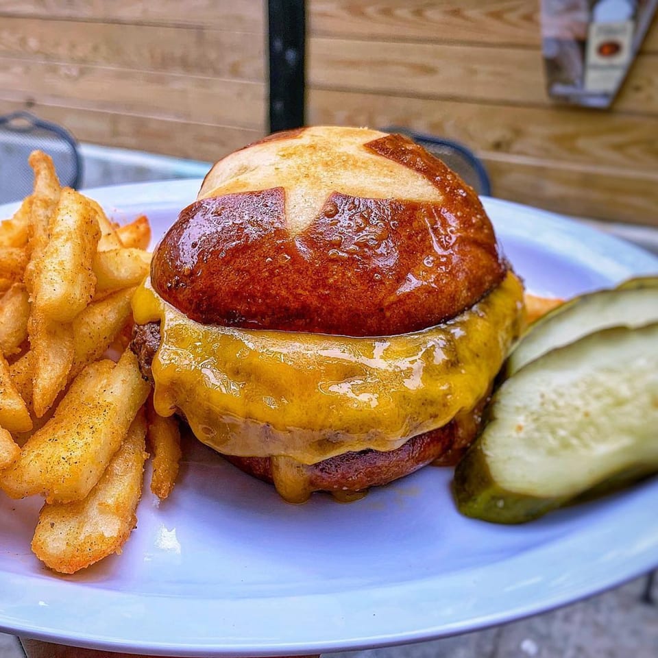 Cheeseburger on a pretzel bun with a side of seasoned fries and pickle slices on a white plate.